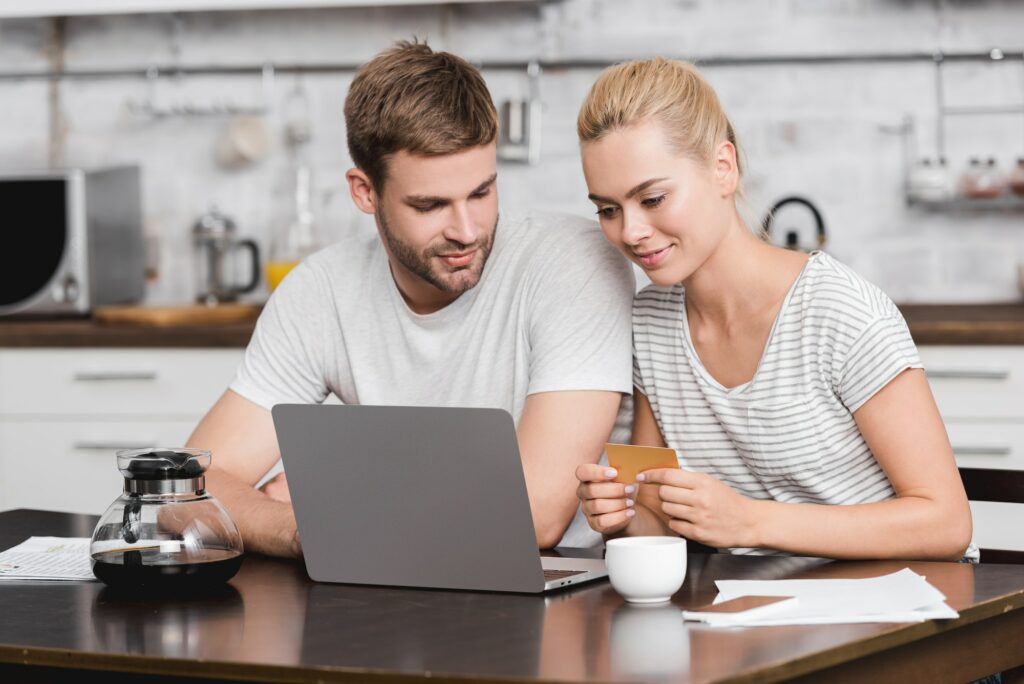 smiling young couple holding business card and using laptop together at kitchen table